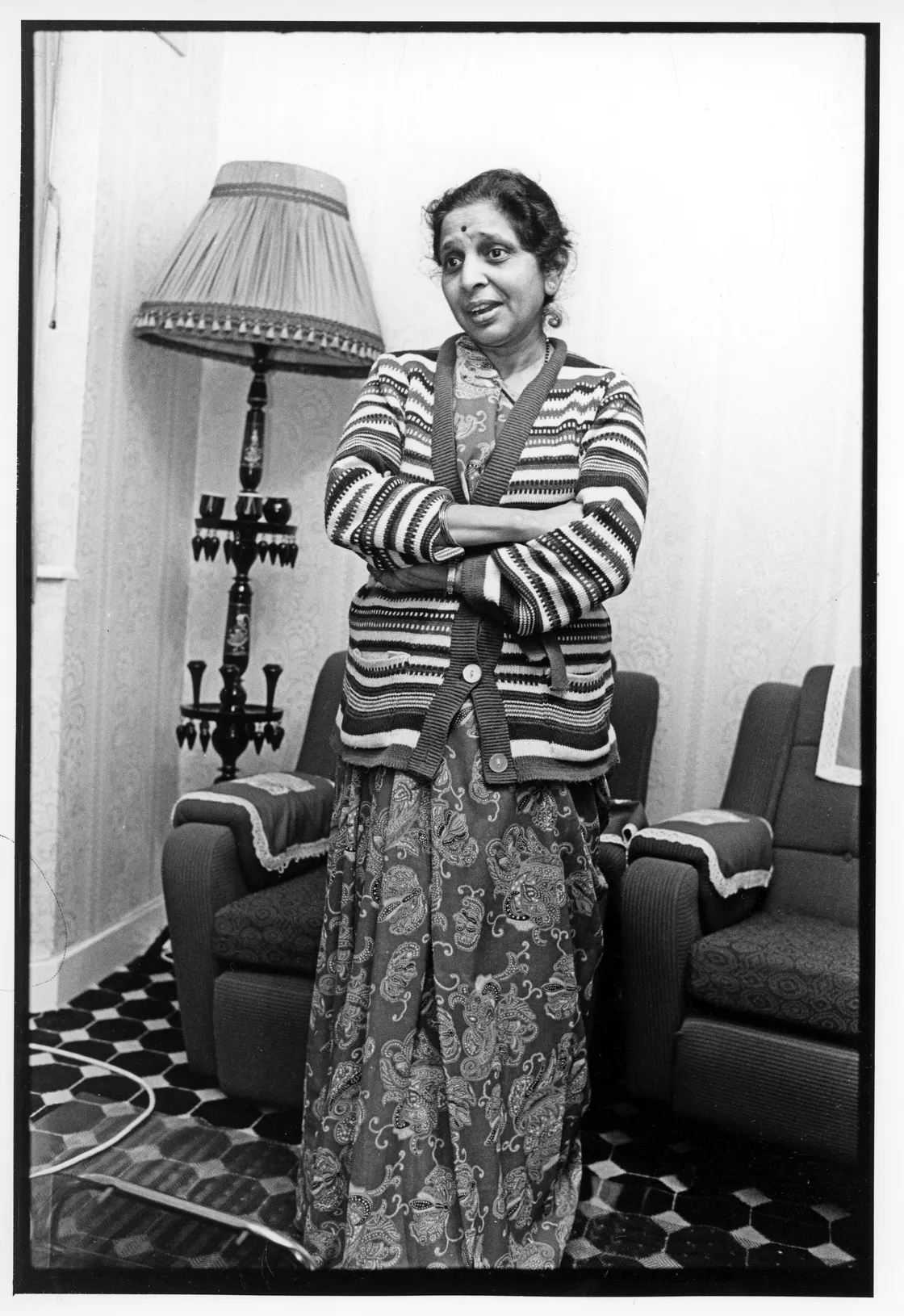 A woman stands in a living room with a patterned carpet and two armchairs, wearing a patterned dress and a striped cardigan, her arms crossed, and a lamp with fringed shade in the background.