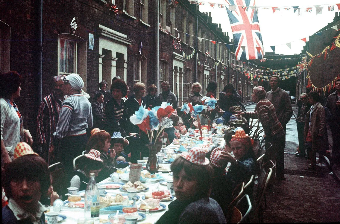 People sat and standing around a long table on a street of terraced housing with decorated with bunting.