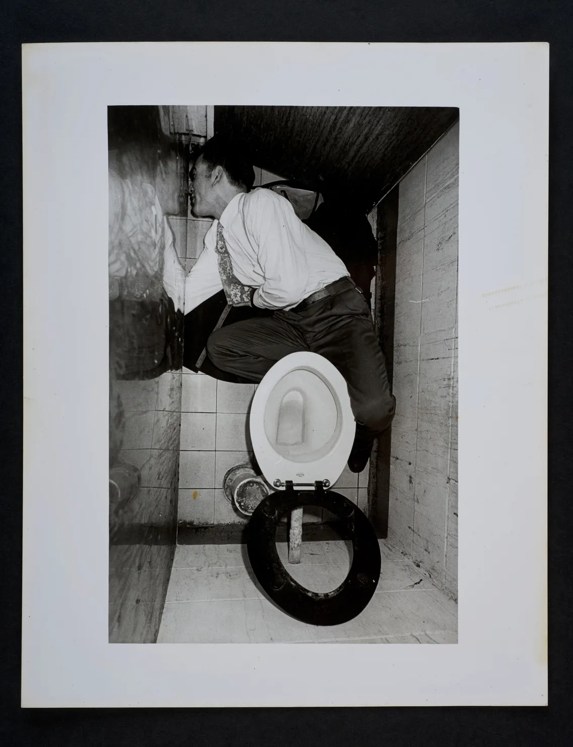 A man in a suit crouches above a toilet in a confined space, peering through a narrow gap in the structure.