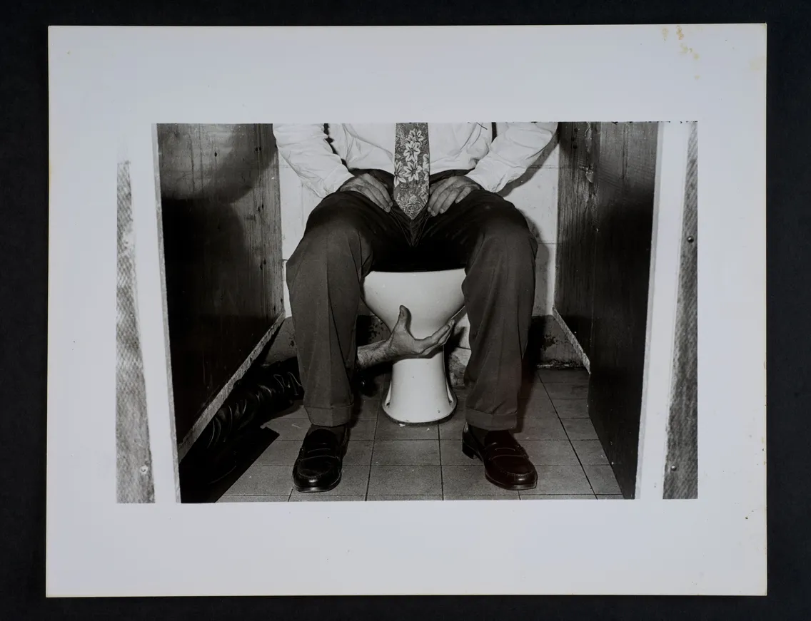 Person sitting on a toilet in a suit and tie, with hands reaching up to grab their knees from between their legs.