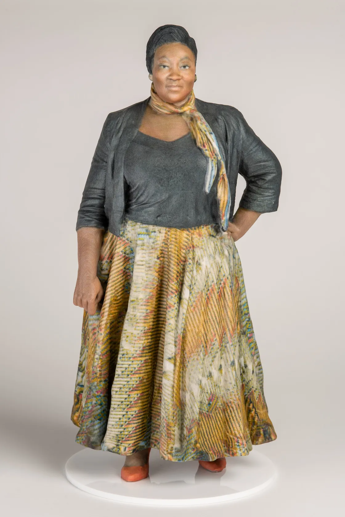 A woman stands facing forward with a neutral expression, wearing a dark top, black jacket, and a patterned skirt. She has one hand on her hip and is standing on a white circular platform.