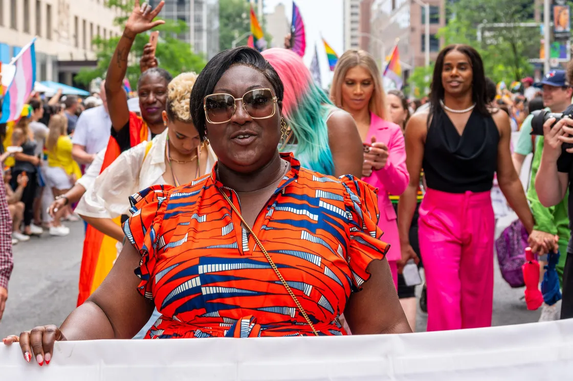 A group of people walk in a parade. The person in front wears large sunglasses and a vibrant red, blue, and orange patterned dress. Colorful flags and a crowd are visible in the background.