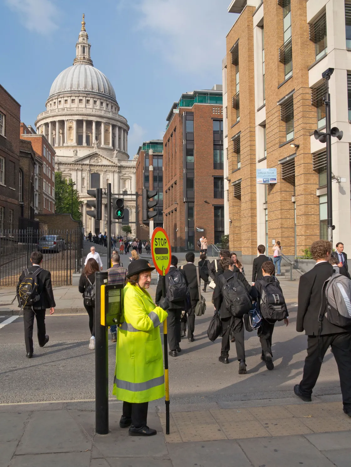 A crossing guard in a bright yellow vest assists pedestrians, including children in school uniforms, to safely cross a street near St. Paul's Cathedral in a cityscape.