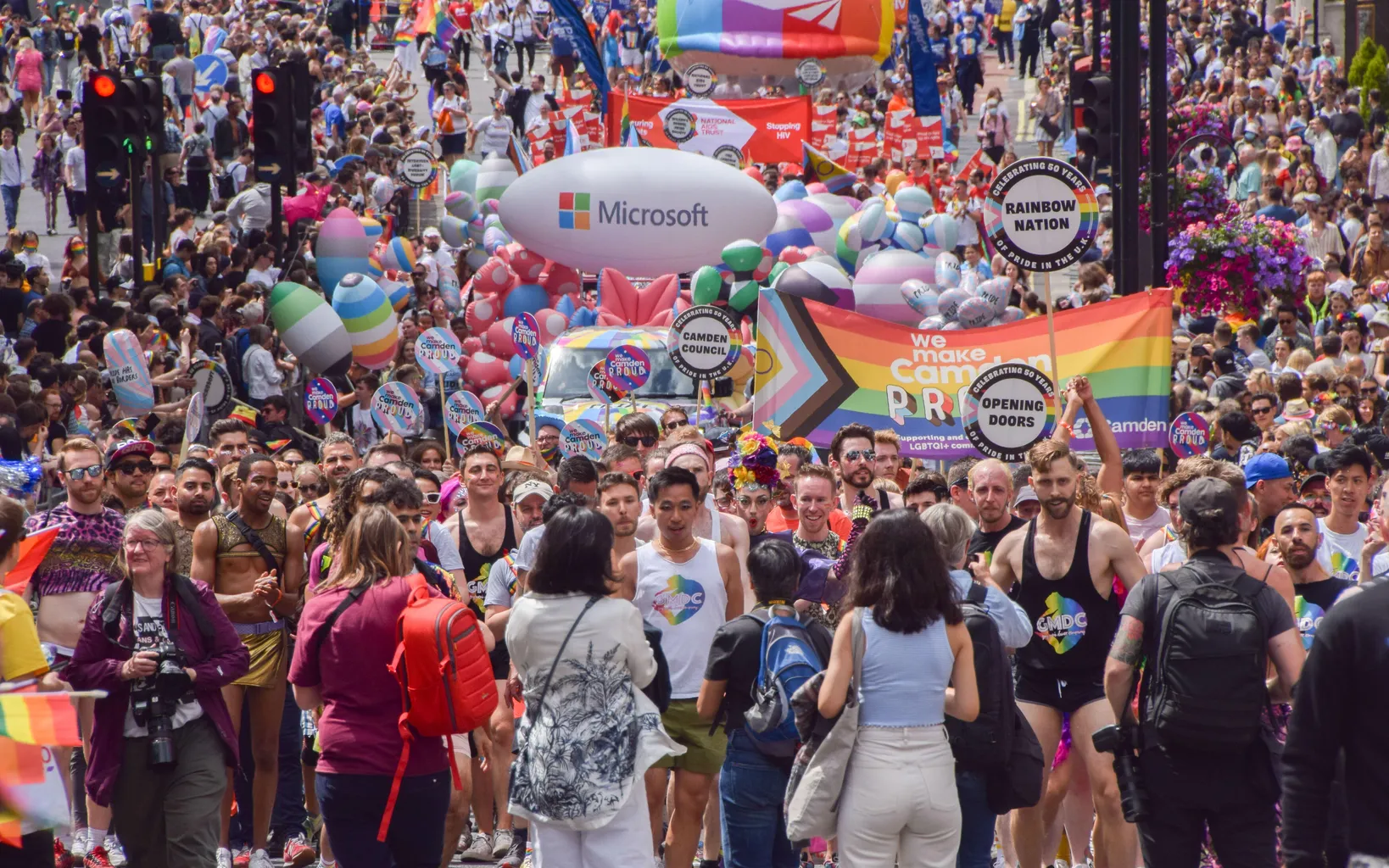 A large crowd participates in a colorful pride parade with various signs and banners, including a prominent Microsoft balloon and a rainbow flag. People are marching down a city street.