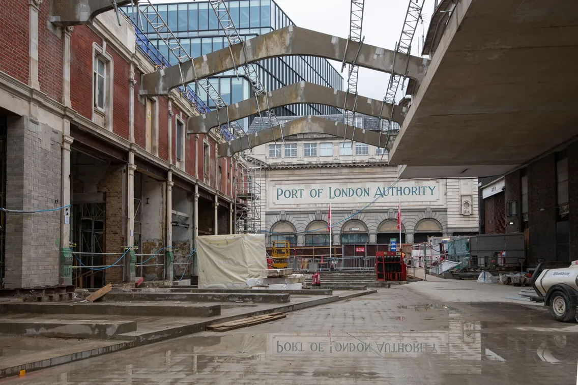 London Museum construction site at Smithfield in Farringdon, unfinished structures and scaffolding ite withstands in front of a building labeled "Port of London Authority." The ground is wet, reflecting parts of the site.