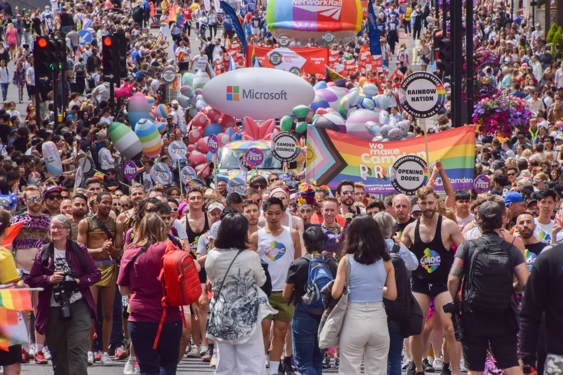 A large crowd participates in a colorful pride parade with various signs and banners, including a prominent Microsoft balloon and a rainbow flag. People are marching down a city street.