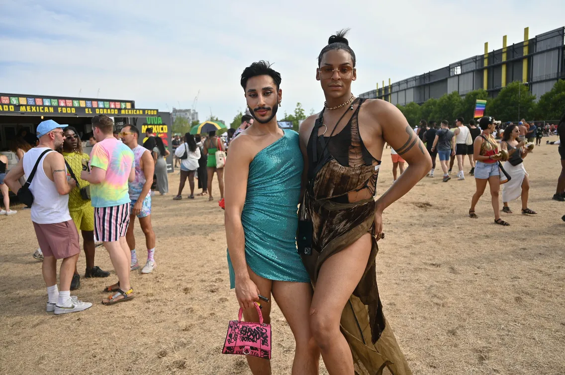 Two people posing outdoors at an event, both wearing fashionable outfits. One is in a shimmery blue dress holding a pink bag, and the other is in a brown patterned dress with accessories. People in the background.