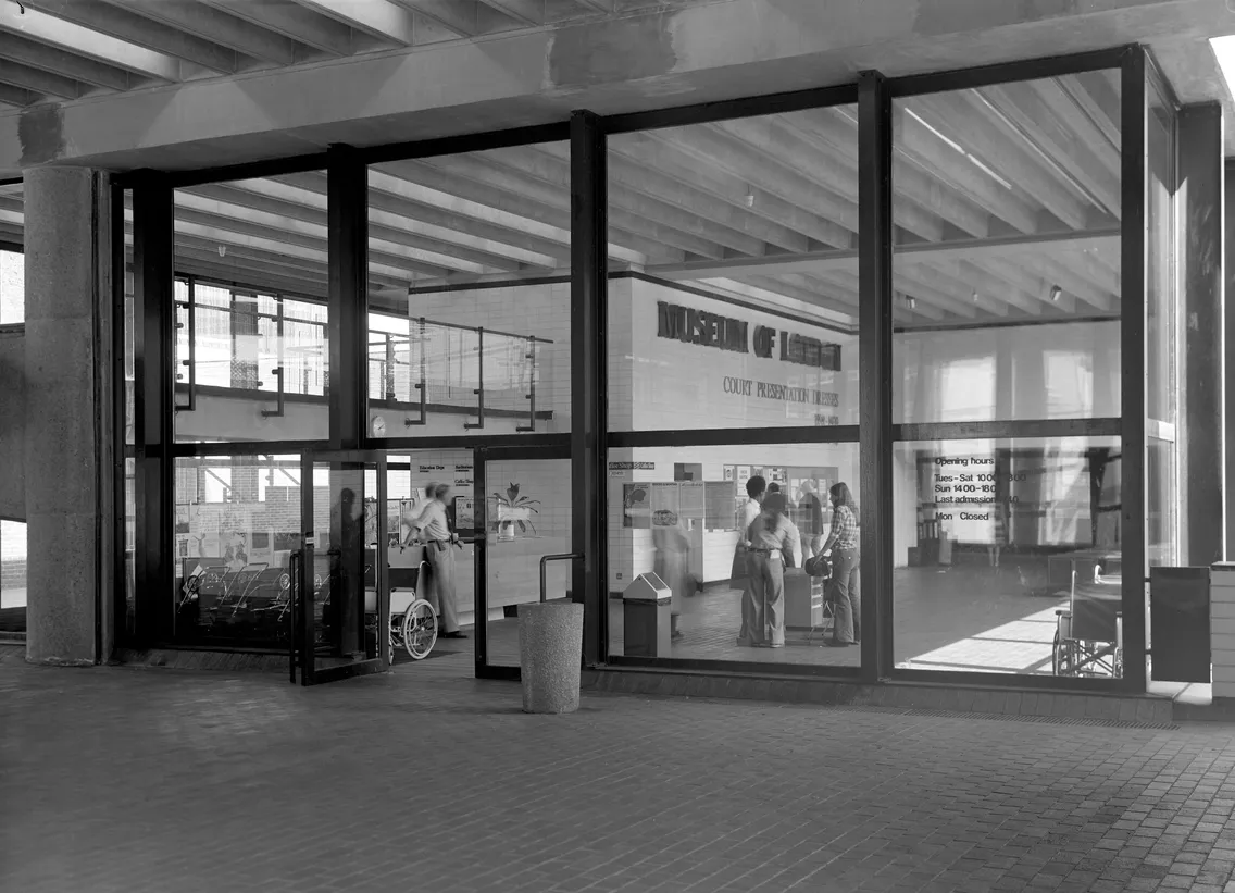 A black-and-white photo of a glass-walled museum exhibit. Visitors are seen inside viewing displays. Large text on the back wall reads "Museum of Minerals." Spaces in the museum are well lit.