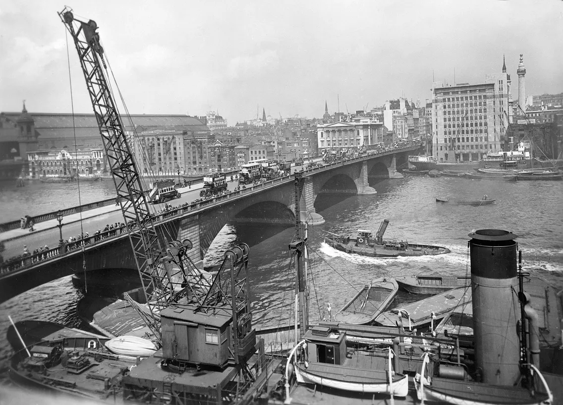 Historic black and white photo of a bustling river port with cranes, moored boats, and a multi-arch bridge spanning the river, with city buildings in the background.