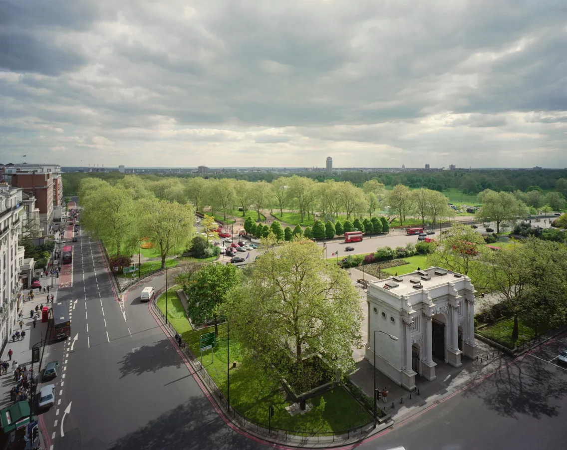 Aerial view of a bustling city intersection with cars and a green park, overlooked by classical-style buildings under a cloudy sky.