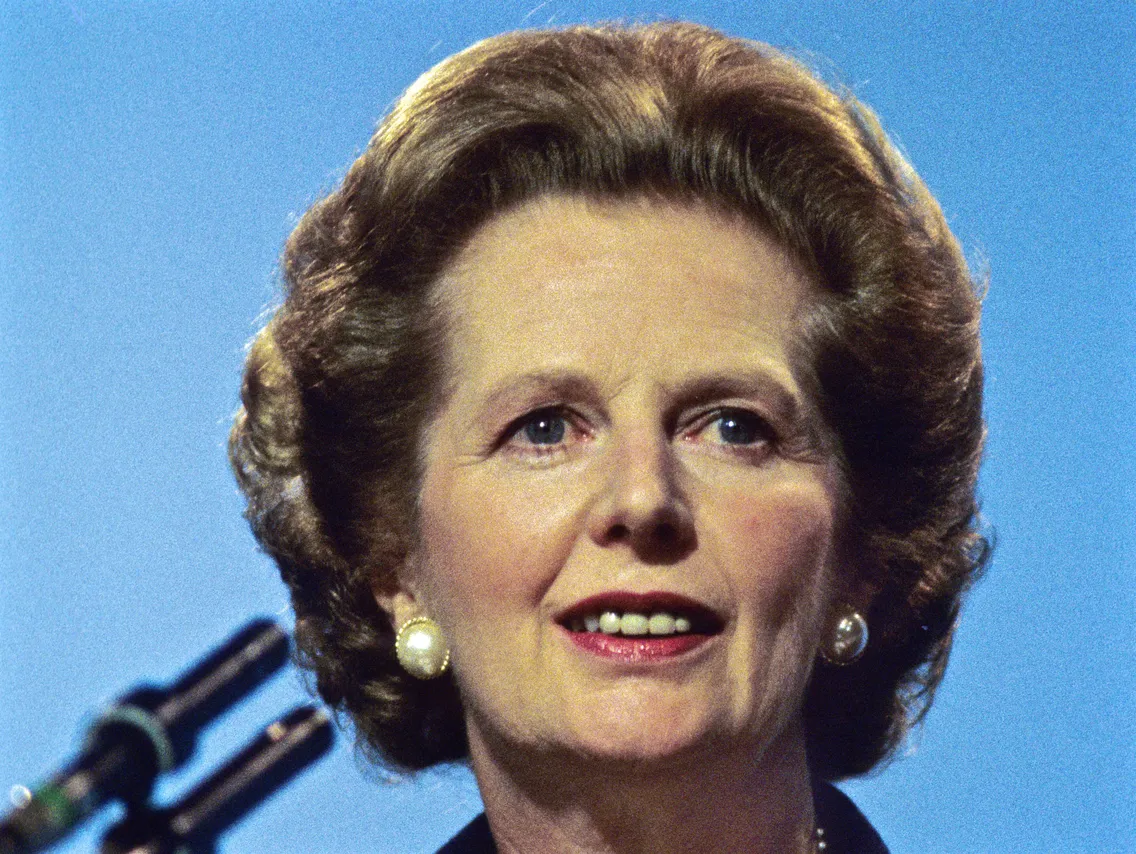 A person with styled hair and wearing pearl earrings is speaking in front of a blue background with a microphone in view.