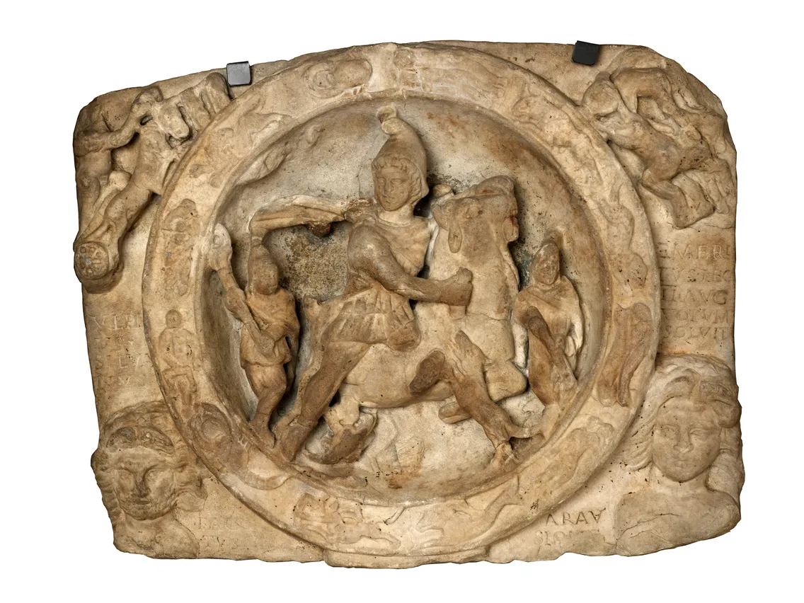Ancient stone relief depicting a warrior on horseback battling a lion, surrounded by decorative motifs and inscriptions.