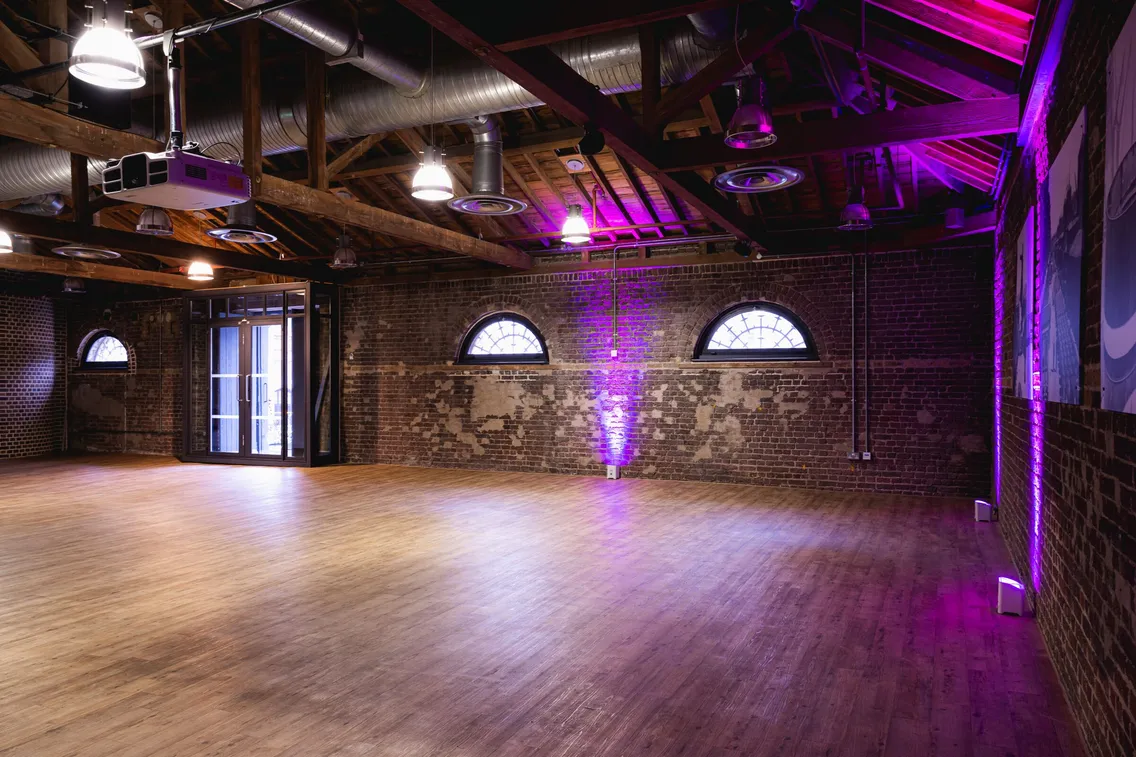 A spacious room with a wooden floor, exposed brick walls, and large ceiling beams. The room is lit with purple lights and features arched windows and industrial-style air ducts.