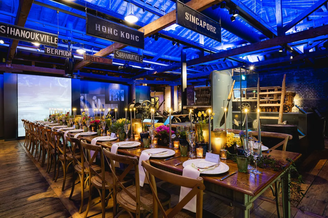 A dining area with long tables and chairs is decorated with candles and flowers under blue lighting, surrounded by signs for various international locations such as Singapore and Hong Kong.