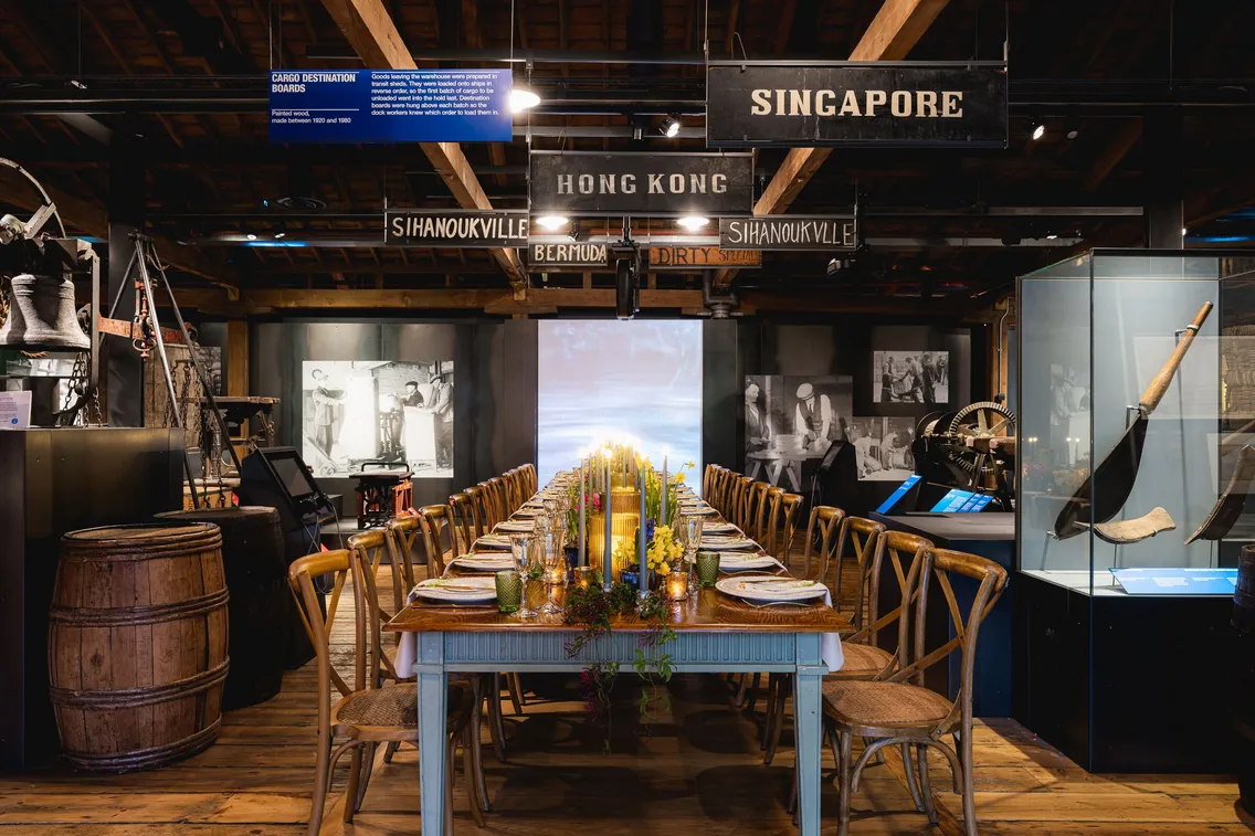 A large dining table set with plates, glasses, and candles is in a rustic room with wooden beams. Overhead signs indicate various cities such as "Singapore" and "Hong Kong.