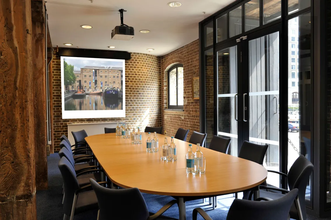 A modern conference room with a wooden table, black chairs, exposed brick walls, and a large window. A projector screen displays an image of a building by a canal. Bottles of water are on the table.