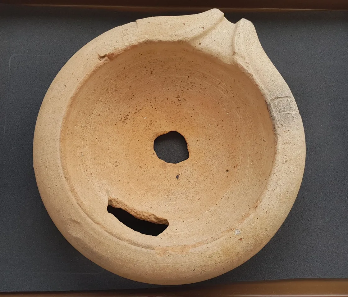 A round, weathered clay bowl with a central hole and a significant crack on one side. The bowl appears to be an artifact or a piece of pottery.