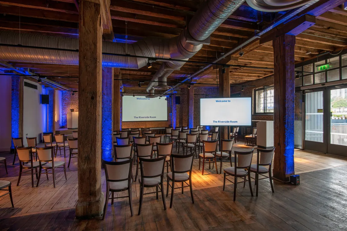 Wooden conference room with exposed beams and ductwork. Rows of chairs face two large screens displaying "Welcome to The Riverside Room." Blue lighting accents the room. A door opens to an outdoor view.