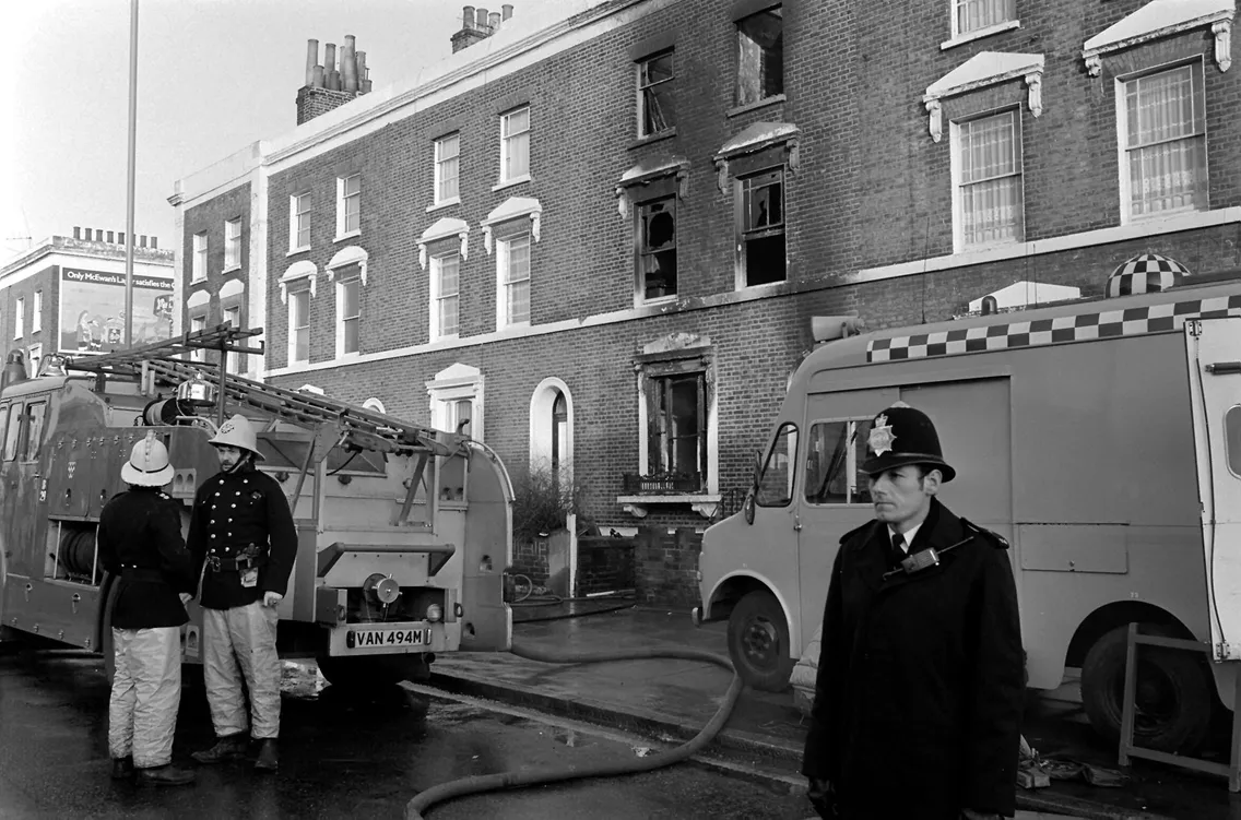 Firefighters and a police officer at the scene of a building fire with fire trucks and hoses in the foreground and a damaged brick building in the background.