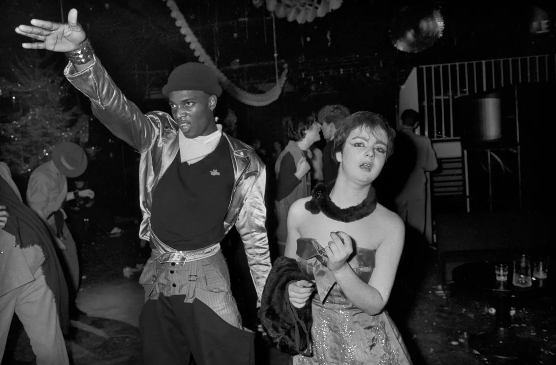 Two people dance in a dimly lit club, one making an energetic gesture. Other attendees are visible in the background. The atmosphere is lively and festive.