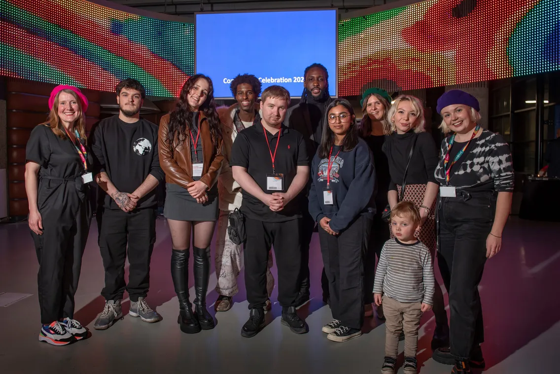 A group of twelve people, including a small child, stand indoors in front of a screen displaying "Celebration 2023." They are casually dressed and looking at the camera.