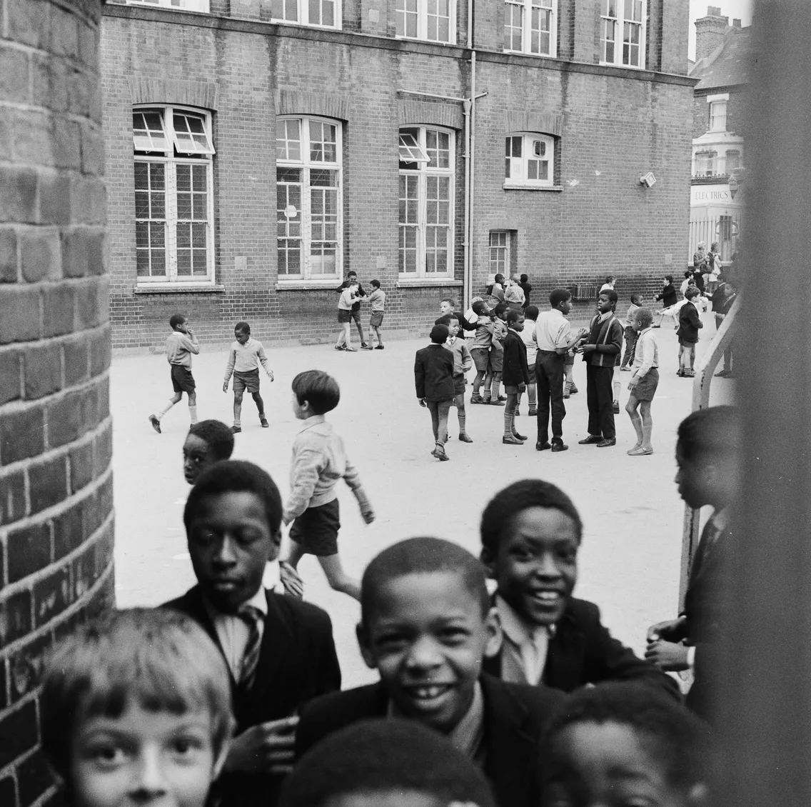 Children of diverse ethnicities playing and smiling in a schoolyard, with a brick school building in the background.