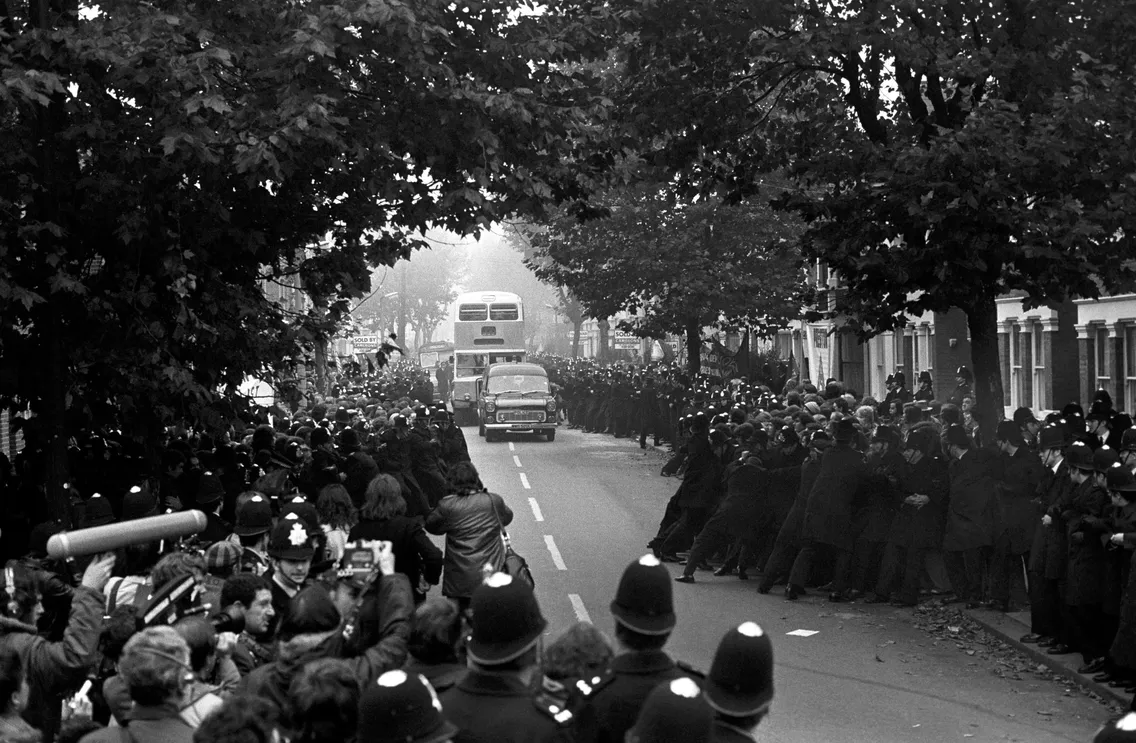 A large crowd and police gather on a street with trees. A double-decker bus and cars are in the background. Photographers and onlookers are present.