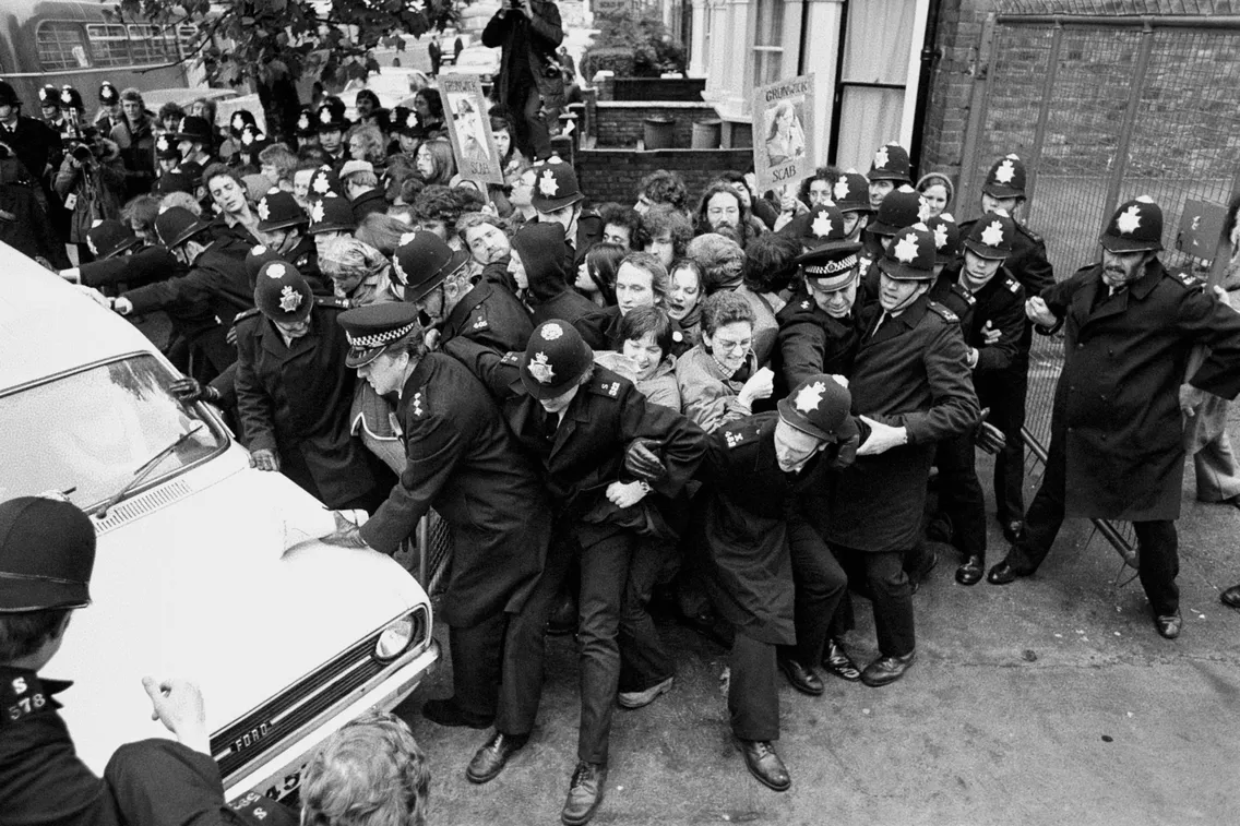 A large group of police officers and civilians struggle near a white vehicle. Some officers hold back civilians while others push forward. The setting appears to be outside a building.