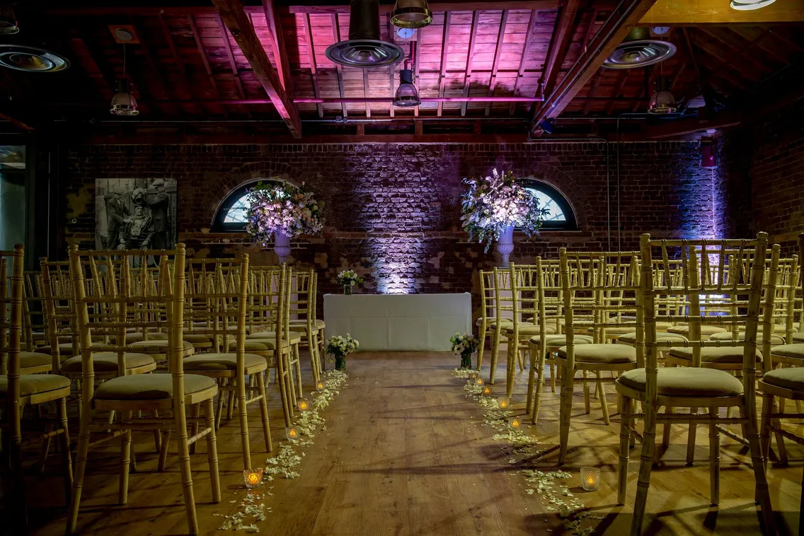 A wedding ceremony setup in a rustic venue with wooden chairs arranged in rows, floral decorations, a petal-lined aisle, and an altar at the front. The room has exposed brick walls and a wooden ceiling.