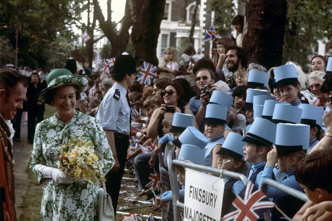 A smile from Queen Elizabeth II as she meets people during a in a park, including a group of children wearing blue hats.