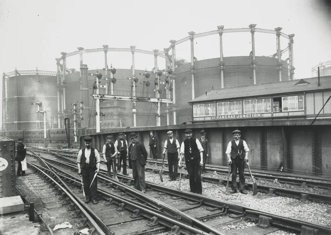 Historical black and white photo of railway workers standing on tracks at St Pancras junction, surrounded by large gasometers and railway infrastructure.