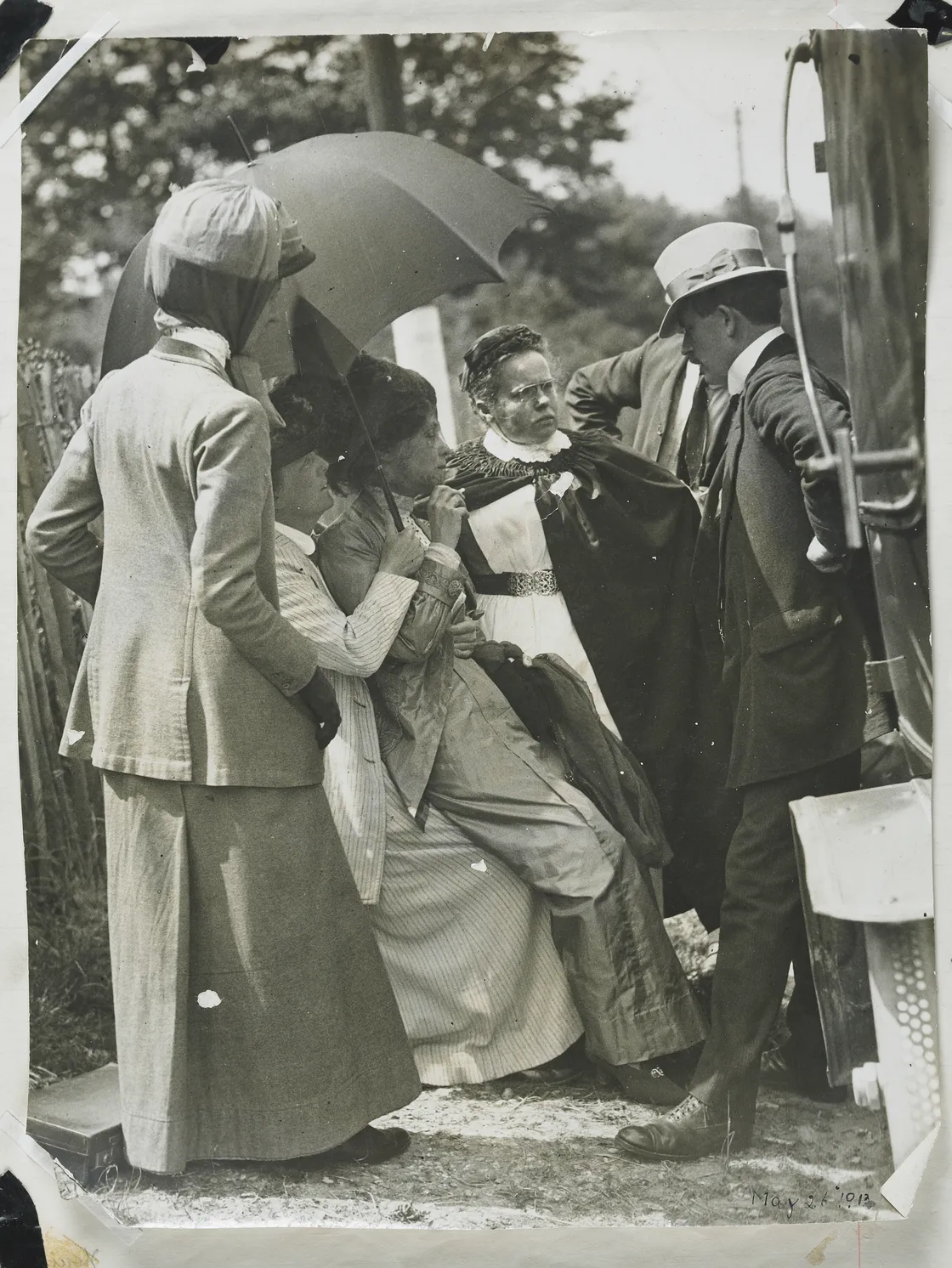 A black and white vintage photo from 1912 showing a group of people gathered under an umbrella, conversing beside an early model car.