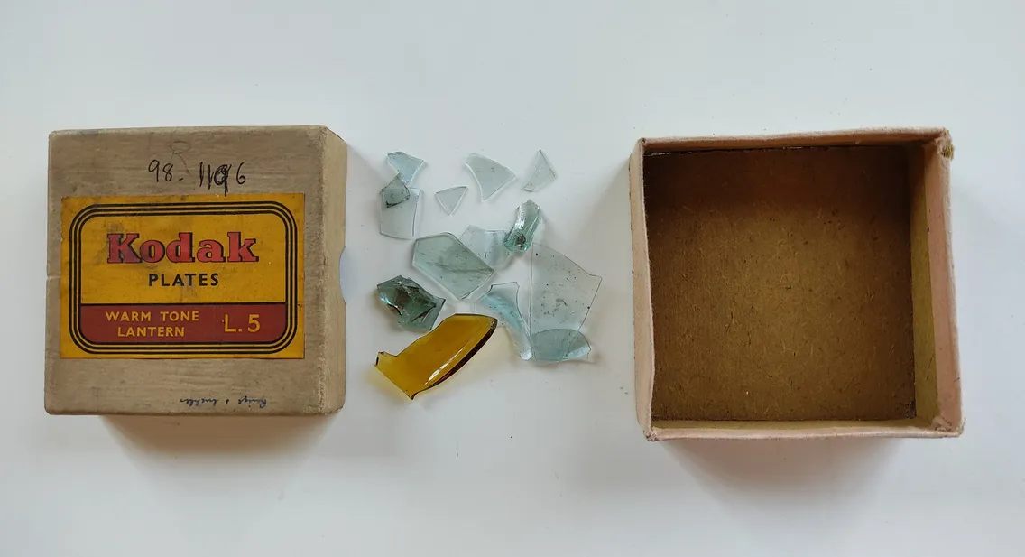 A Kodak Plates box labeled "Warm Tone Lantern L5," an assortment of broken glass pieces beside it, and an open, empty box next to the glass.