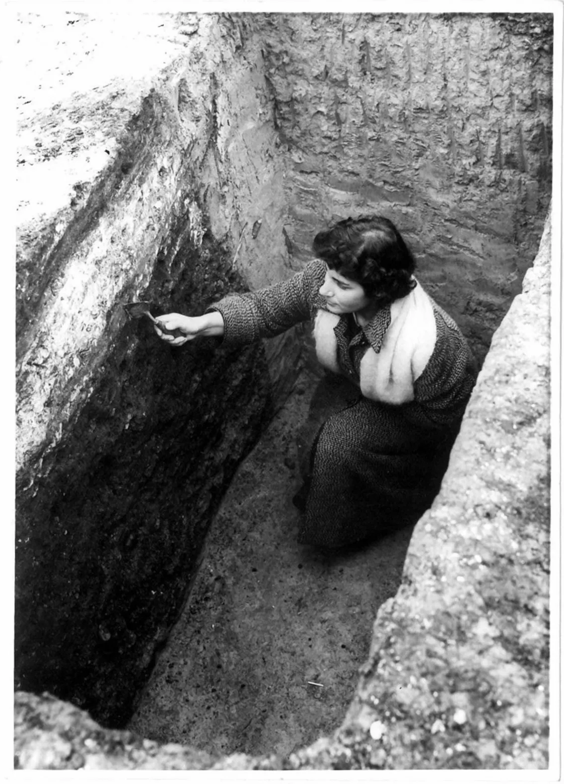 A woman in a coat kneels in an excavated pit, carefully examining and scraping the wall with a tool.