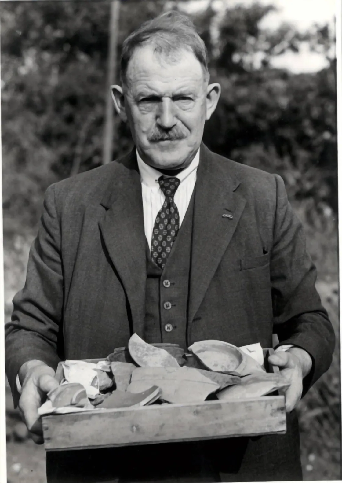 A man in a suit and tie holds a tray filled with various pottery fragments or pieces, standing outdoors.
