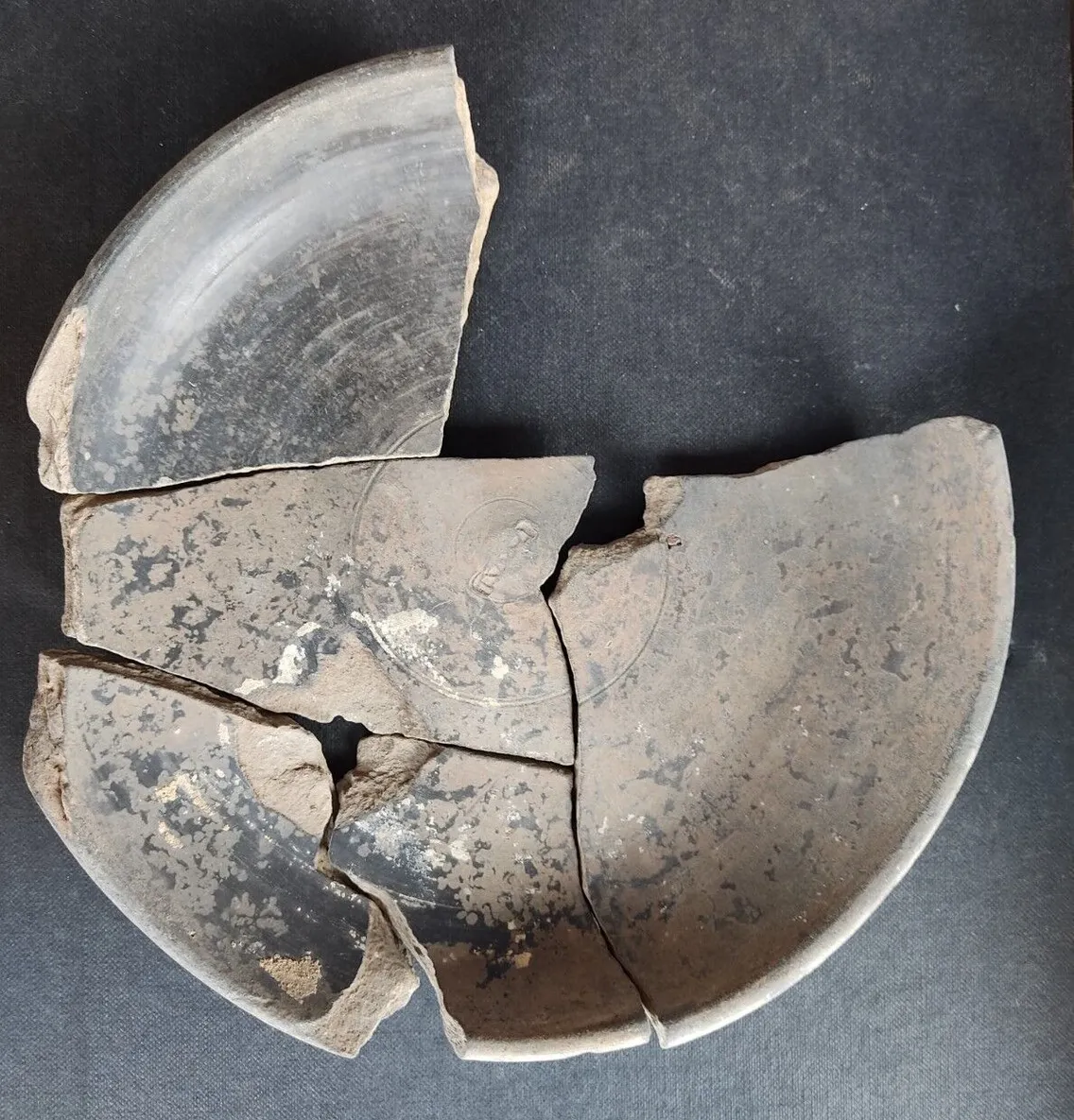 A partially reassembled clay bowl with several broken and missing pieces, displayed on a dark surface.