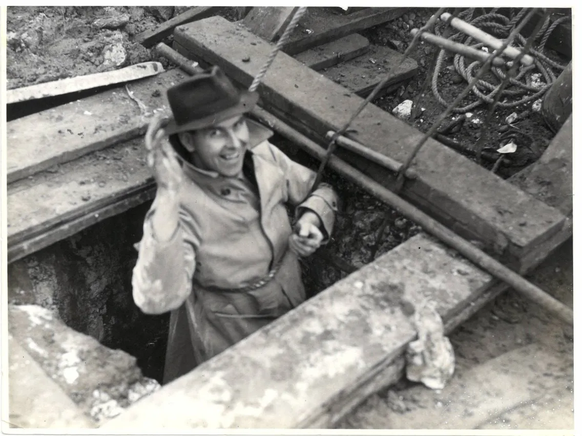 A person in a trench coat and hat is coming out of a manhole or underground passage, with a smile and holding onto metal bars. The area around is unfinished or under construction.