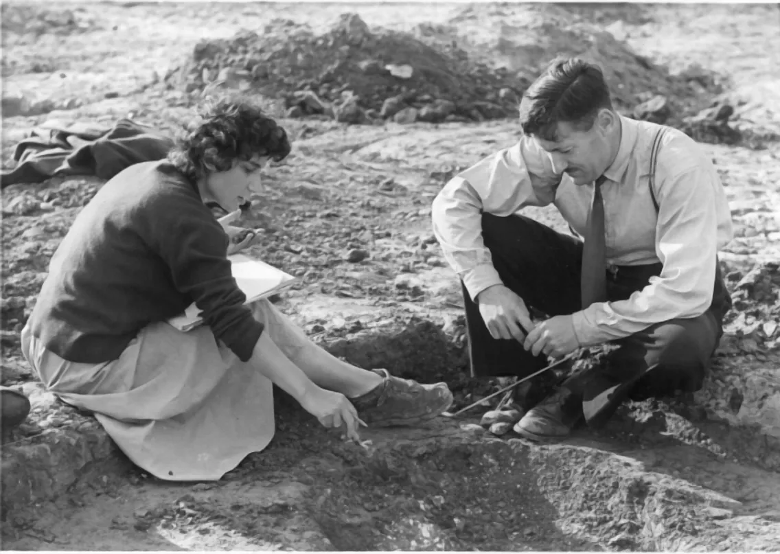 Two people, one holding a notebook and the other using a tool, examine an archaeological dig site.
