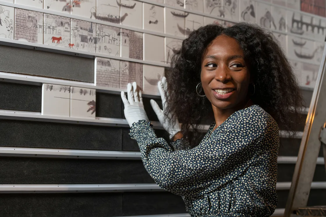 A black woman wearing white gloves holding tiles and looking away from the camera