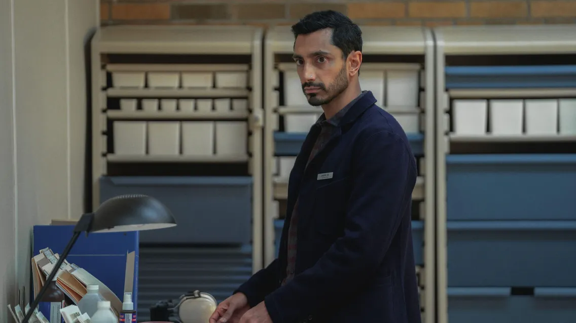 A man with short black hair and a beard stands in an office, wearing a dark jacket and facing a desk with office supplies and a lamp. Shelves are visible in the background.