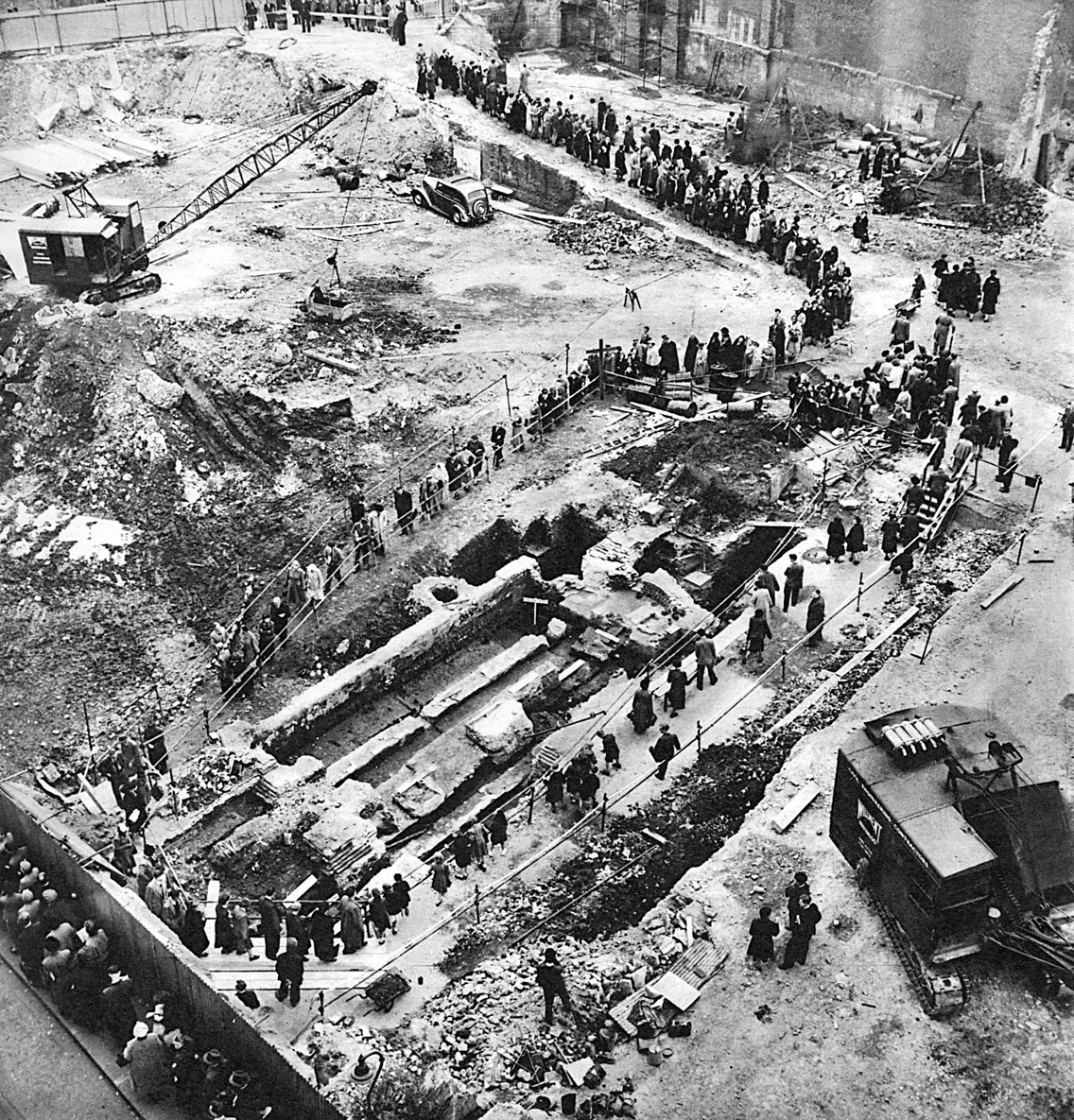 A large crowd gathers to observe an archaeological excavation at a construction site, where the foundation of an ancient structure is visible. Construction equipment and vehicles are present in the background.