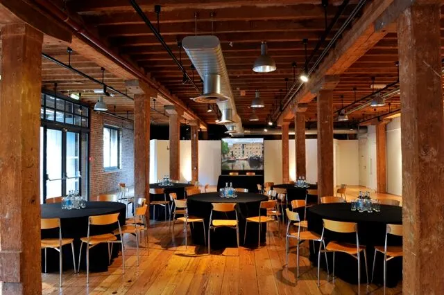 A spacious industrial-style event room with wooden beams, round tables covered in black tablecloths, and chairs arranged around, prepared for a gathering or conference.