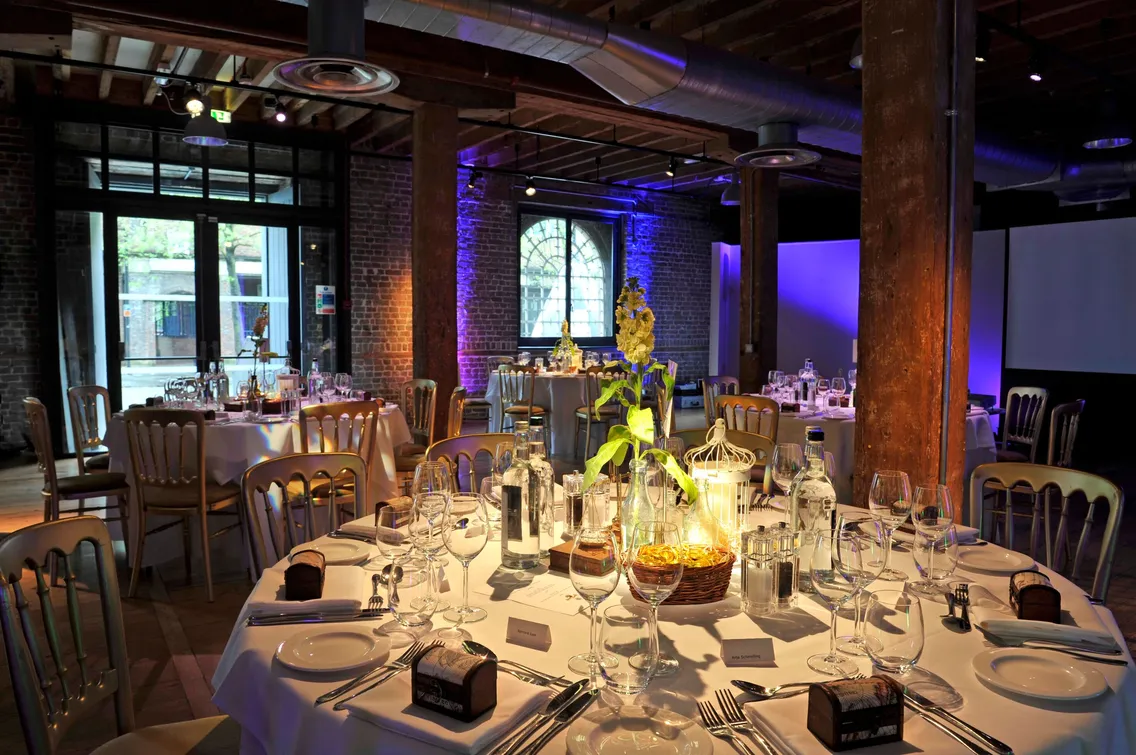A dimly lit, elegant dining hall with round tables set for a formal event. Each table features place settings, glassware, and decorative centerpieces. Exposed brick walls and wooden beams create a rustic ambiance.