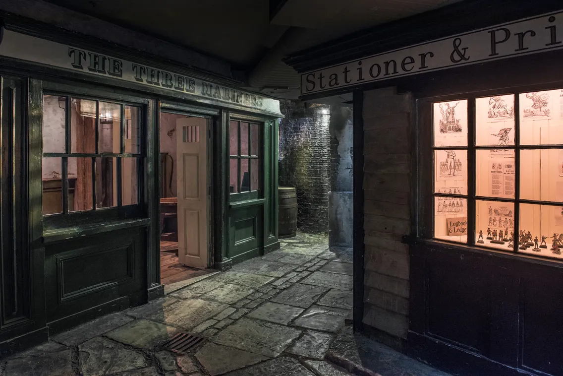 A dimly lit alley with old-fashioned storefronts, including a pub named "The Three Mariners" and a stationer & printer shop with illuminated windows displaying various posters.