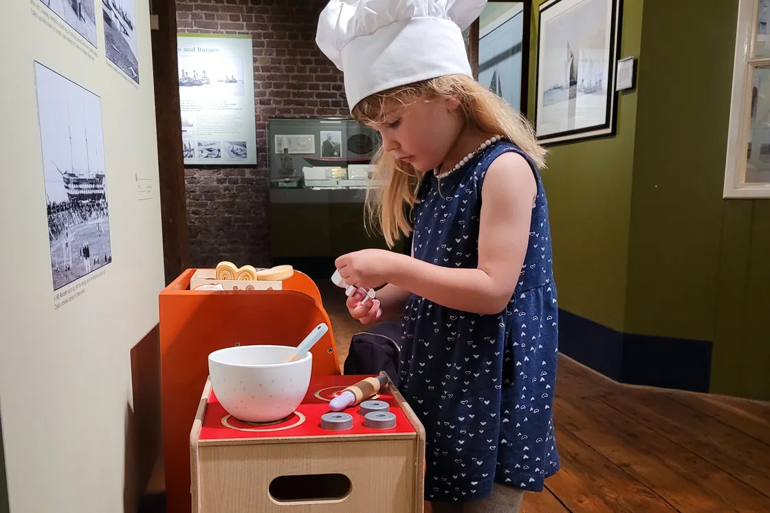 Two girls in a recreation of an old-style, one stood wearing a hairnet and one sat down holding a mug