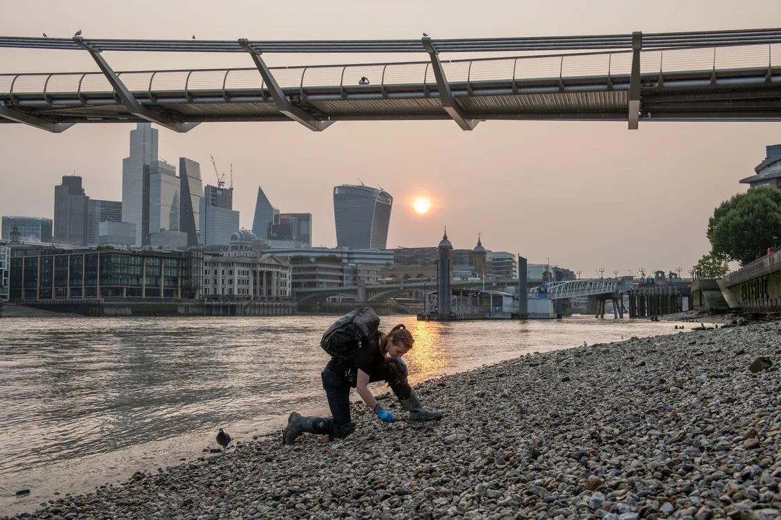 A person is crouched on a pebbled riverbank near a body of water under a footbridge, with a skyline of modern skyscrapers visible in the background during sunset.