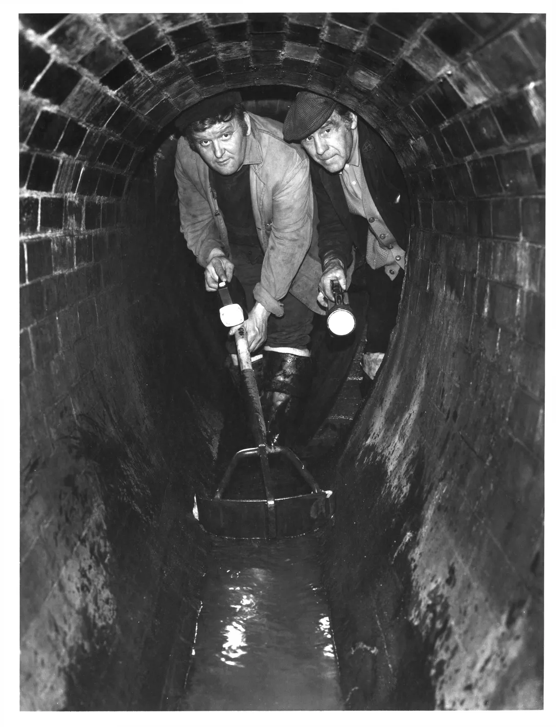 Two men examining water quality in a sewer tunnel, one holding a flashlight and the other a sampling container.