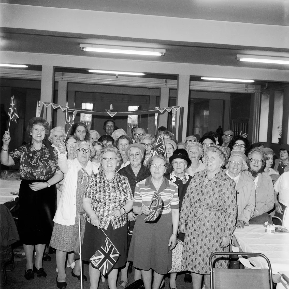 A group of elderly women holding small flags, smiling at a social gathering in a decorated hall.