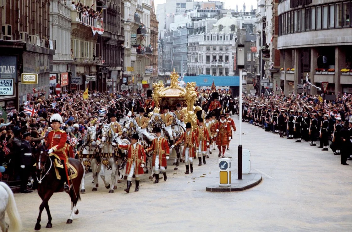 The Golden State Coach with Queen Elizabeth II and Duke of Edinburgh riding in a carriage through a street led by people in regalia on horses.
