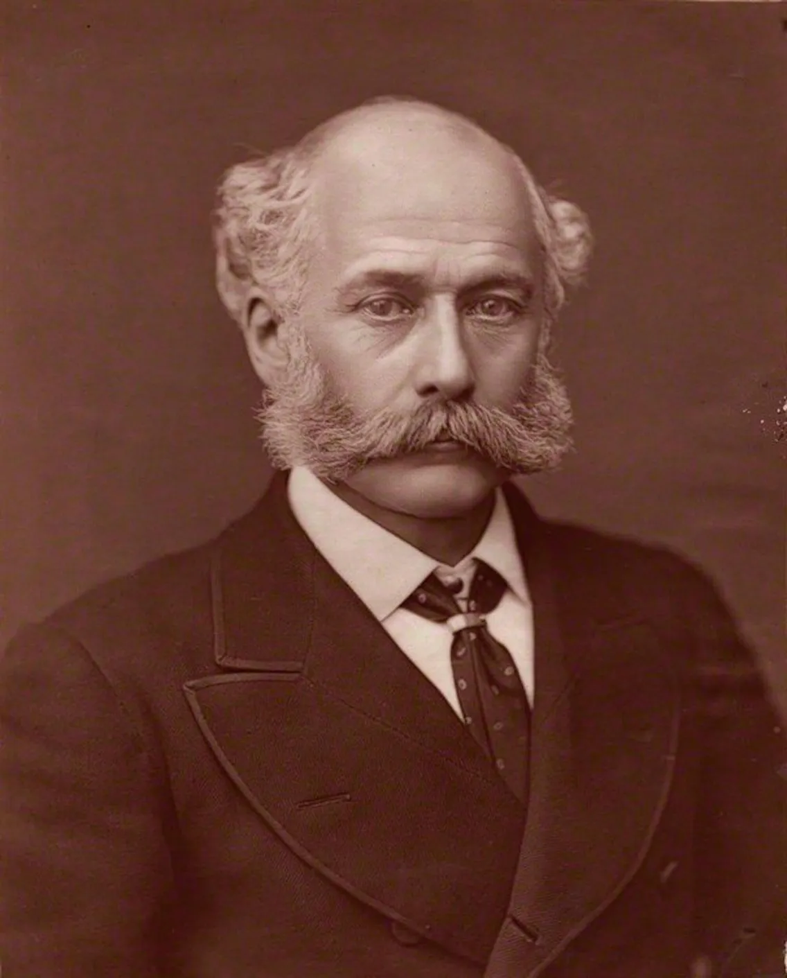 A stern-faced man with bushy sideburns, wearing a suit and tie, poses for a portrait against a plain background.